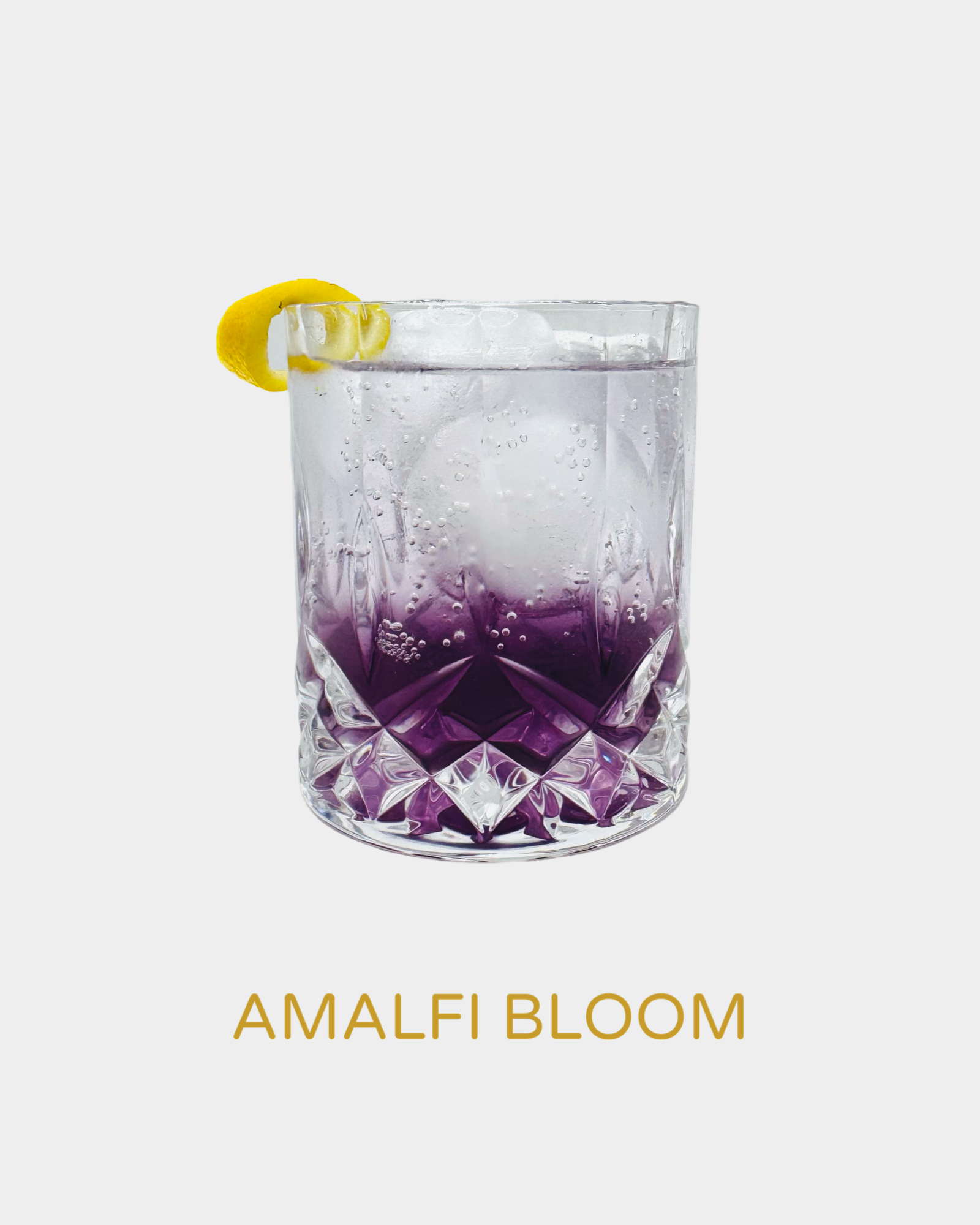 Amalfi Bloom cocktail in a crystal glass with a purple gradient hue, large ice sphere, and lemon twist garnish
